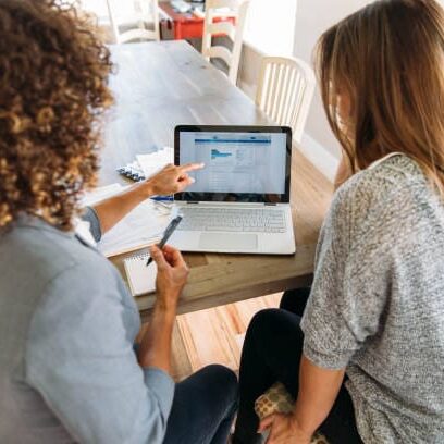 A female financial advisor sits with a woman at her dining room table with laptop and financial reports helping her monthly budget and investments. She is showing her client how to read a financial report as she works on her computer to do monthly finances, pay taxes and save money for the future.